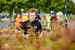 Een jongen met een vastberaden blik rent door een zonnig park met struiken op de voorgrond en deelnemers op de achtergrond tijdens een looponderdeel van de challenge.