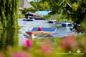 Een zonneboot met startnummer 79 vaart door een smalle watergang, geflankeerd door groen en bloemen, gevolgd door andere boten in wedstrijdformatie.