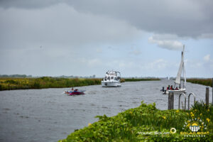 Een zonneboot en een zeilboot passeren elkaar op een open Friese vaarroute, omgeven door riet en wolkenlucht.