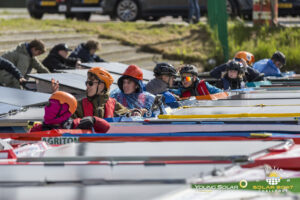 Jongeren met helmen, zonnebrillen en reddingsvesten zitten in hun zonneboten aan de kant van het water en wachten op het startsignaal tijdens de Solar Boat Challenge.