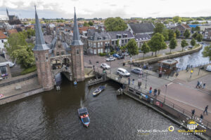 Twee zonneboten varen onder de iconische Waterpoort in Sneek tijdens de Young Solar Challenge, terwijl toeschouwers vanaf de brug en kade toekijken.